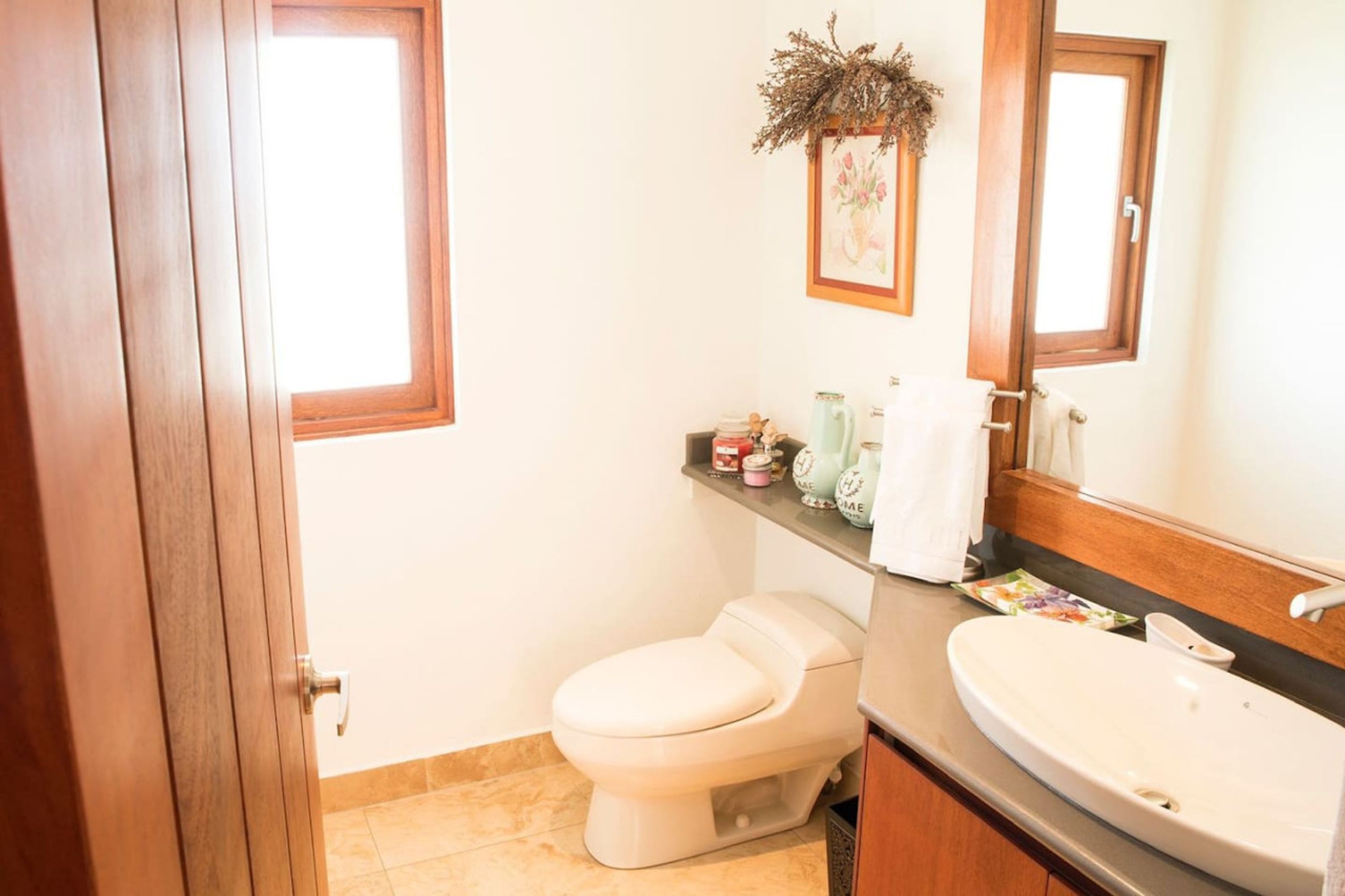 A bathroom with a toilet, sink, and a mirror. There is a shelf with toiletries and a decorative plant.