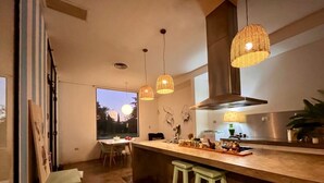 Μια μοντέρνα κουζίνα με μεγάλο κεντρικό πάγκο, κρεμαστά φωτιστικά και θέα προς τον εξωτερικό χώρο.