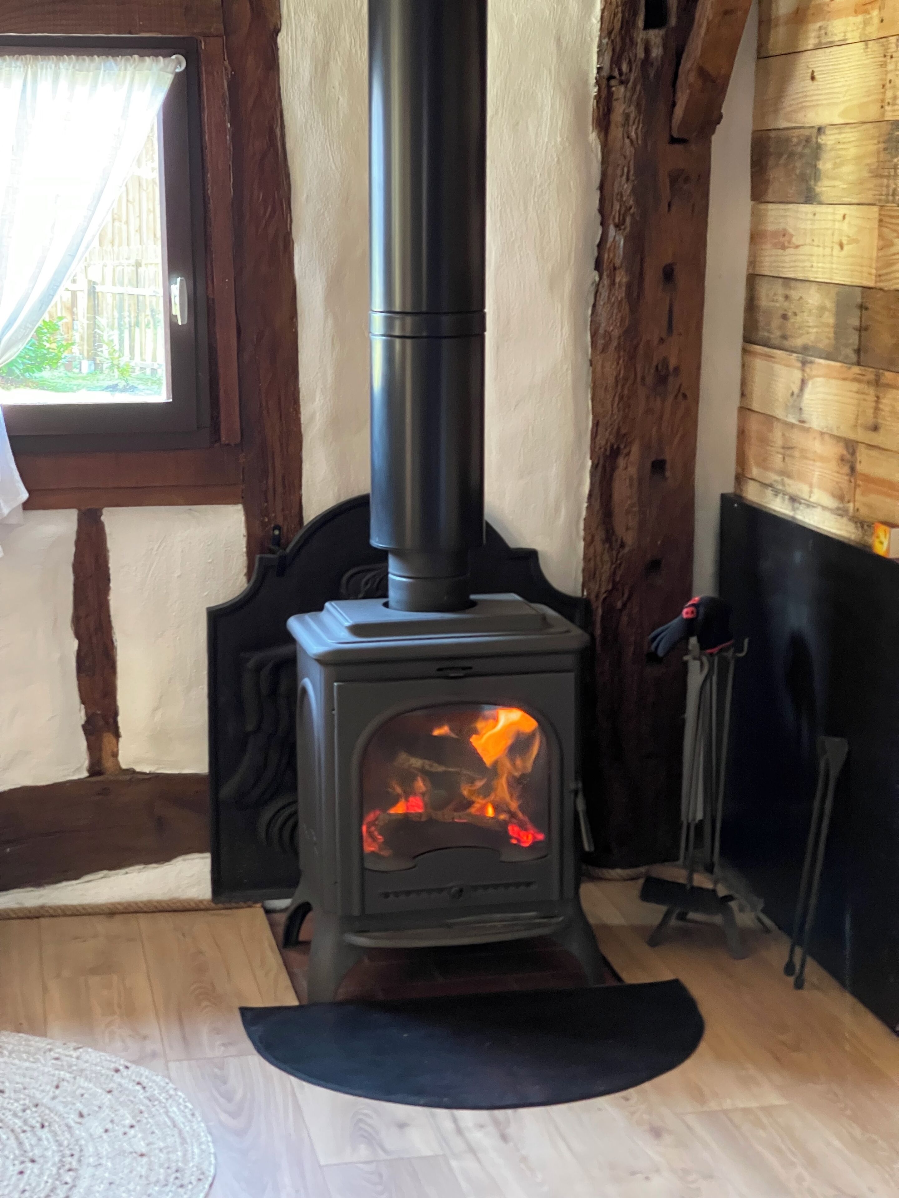 Ein Kaminofen mit sichtbaren Flammen, platziert in einem Raum mit Holzwänden und einem Fenster mit Vorhängen.