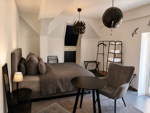Un dormitorio moderno con cama, una silla, una mesa, una lámpara, una lámpara colgante y una ventana.