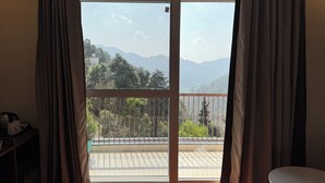 Ένα δωμάτιο με μια μεγάλη συρόμενη γυάλινη πόρτα που οδηγεί σε ένα μπαλκόνι με θέα στα βουνά και τα δέντρα.
