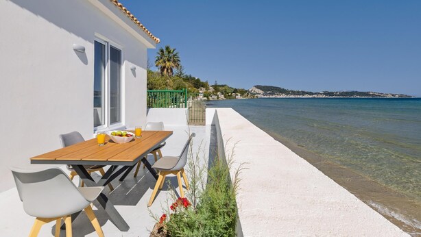 A seaside property with a wooden table and chairs, a small garden, and a view of the sea and distant hills.