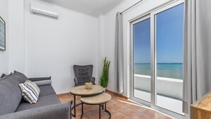 A modern living room with a grey sofa, a small round table, and a chair. There is a large window with a view of the sea.