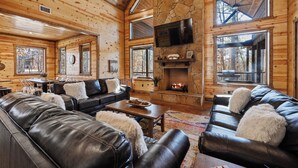 A log cabin interior with a stone fireplace, leather sofas, and large windows.