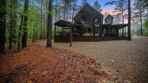 A cabin with a covered porch, surrounded by tall trees and a gravel driveway.