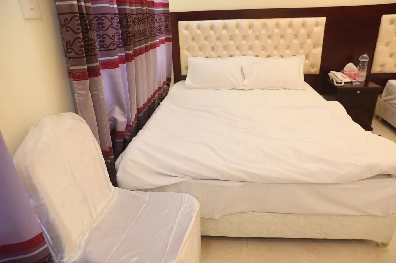 Een hotelkamer met een groot bed, een nachtkastje met een telefoon en zakdoekjes, en een stoel met een witte hoes.