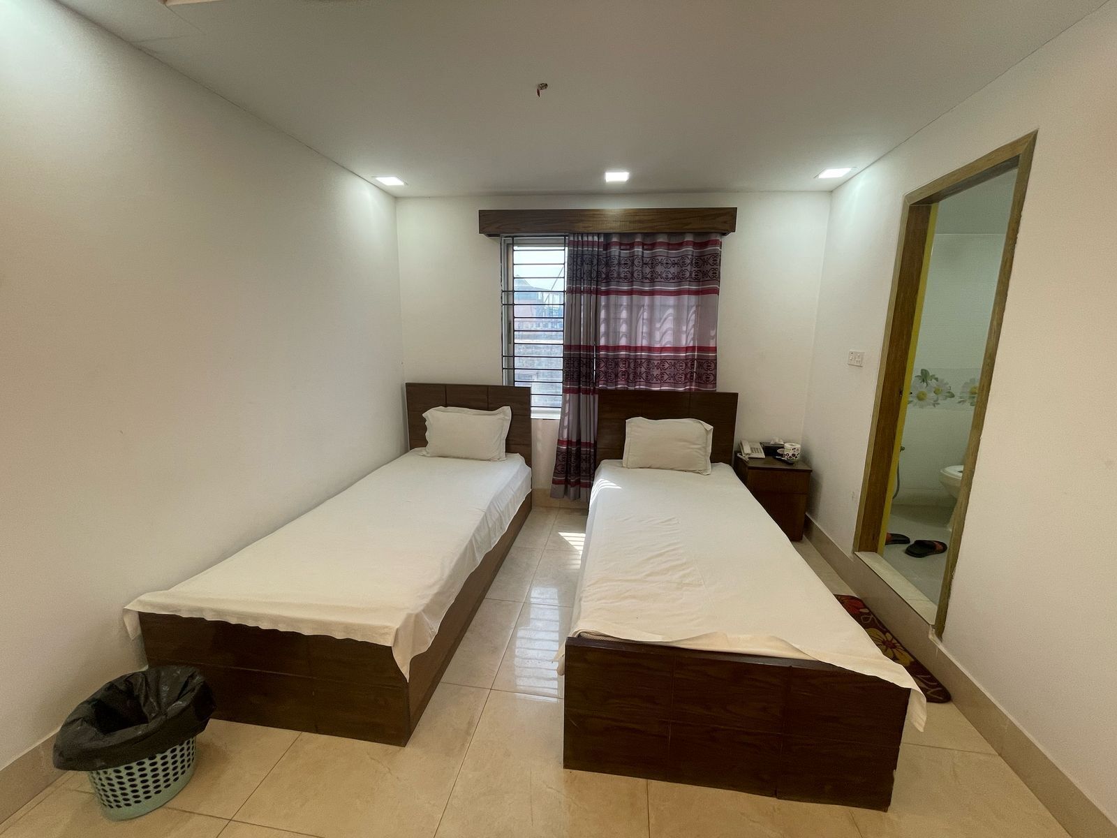 A small, clean room with two single beds, a small trash bin, and a mirror on the right wall.