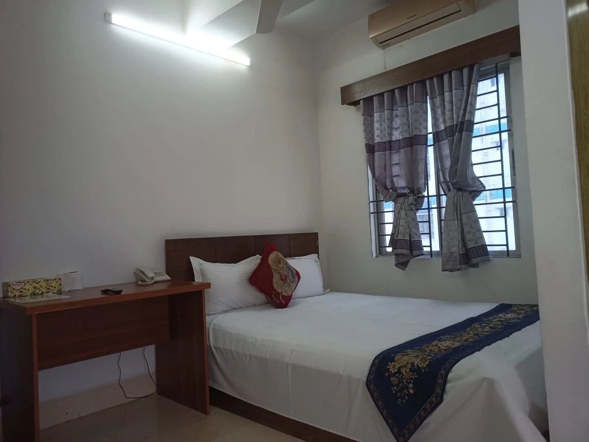 A bedroom with a bed, a desk, a window with curtains, and an air conditioning unit.