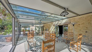 Une vaste terrasse dotée de tables en verre et de chaises en bois, d’un mur en pierre et offrant une vue sur l’extérieur.