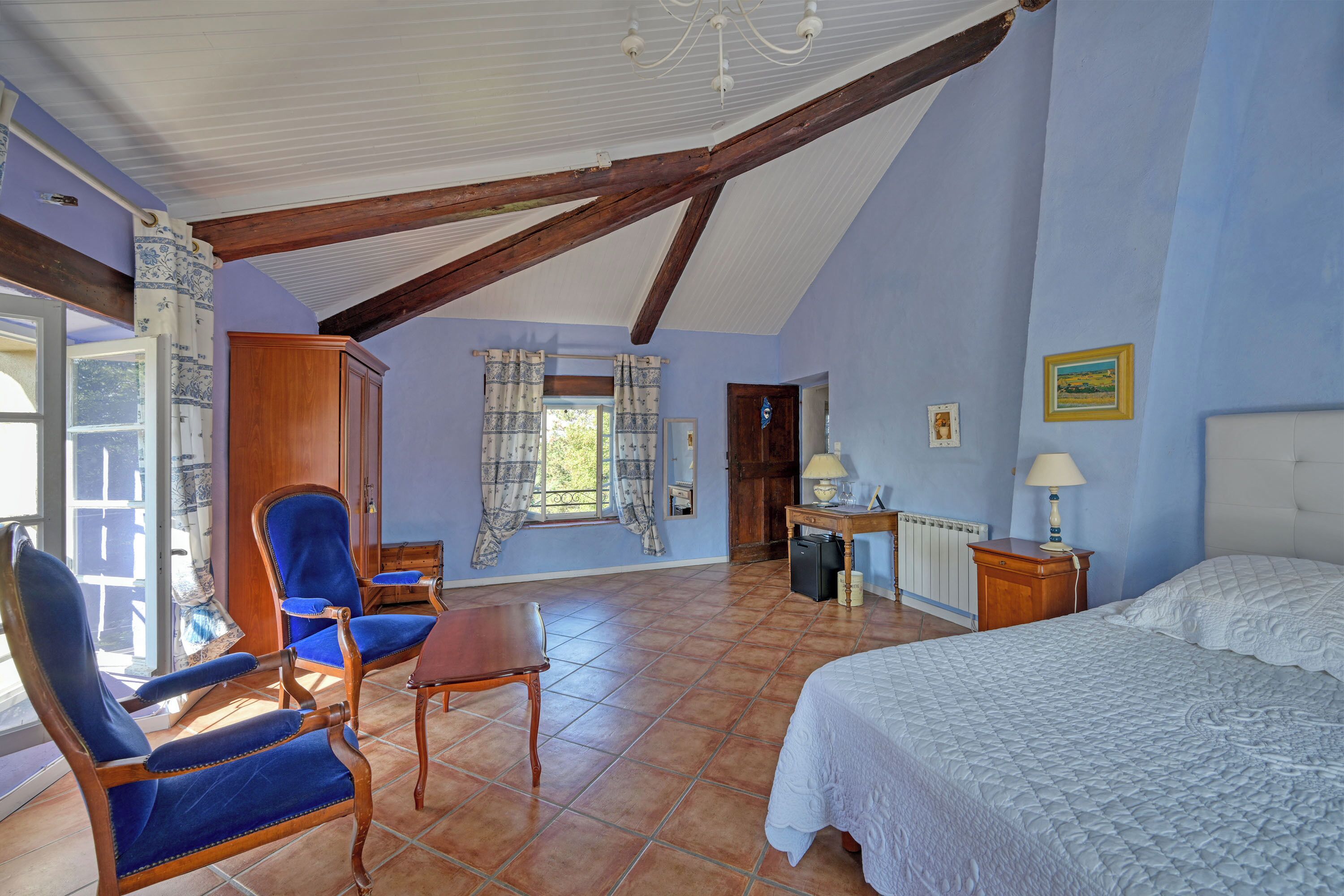 A bedroom with a bed, a chair, a table, a desk, and a window with curtains.