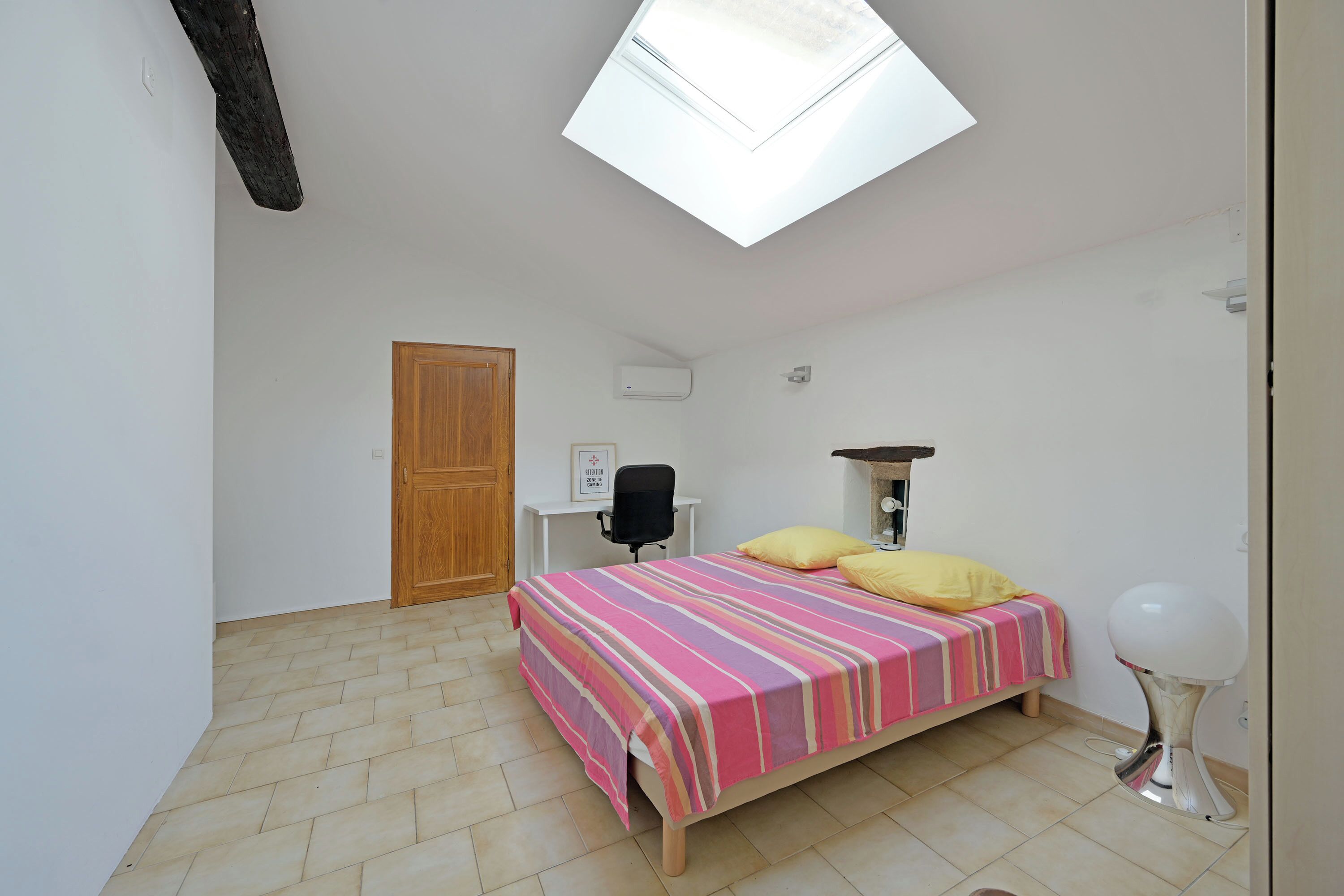 A bedroom with a skylight, a bed with a striped bedspread, a desk with a chair, and a bedside table with a lamp.