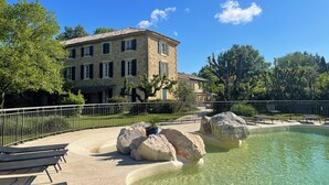 Un bâtiment en pierre avec un toit en tuiles, une piscine à l’eau claire et un espace dallé avec des chaises longues.