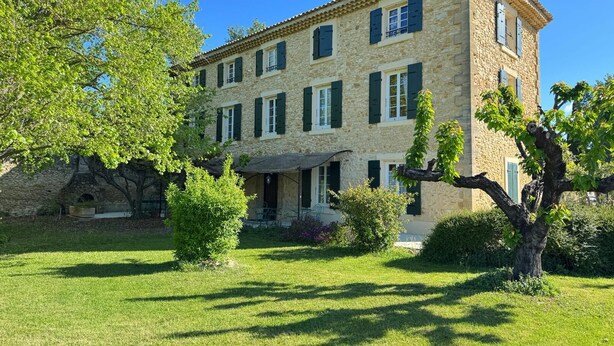 Un bâtiment en pierre doté de nombreuses fenêtres et volets, entouré d’une pelouse et d’arbres bien entretenus.