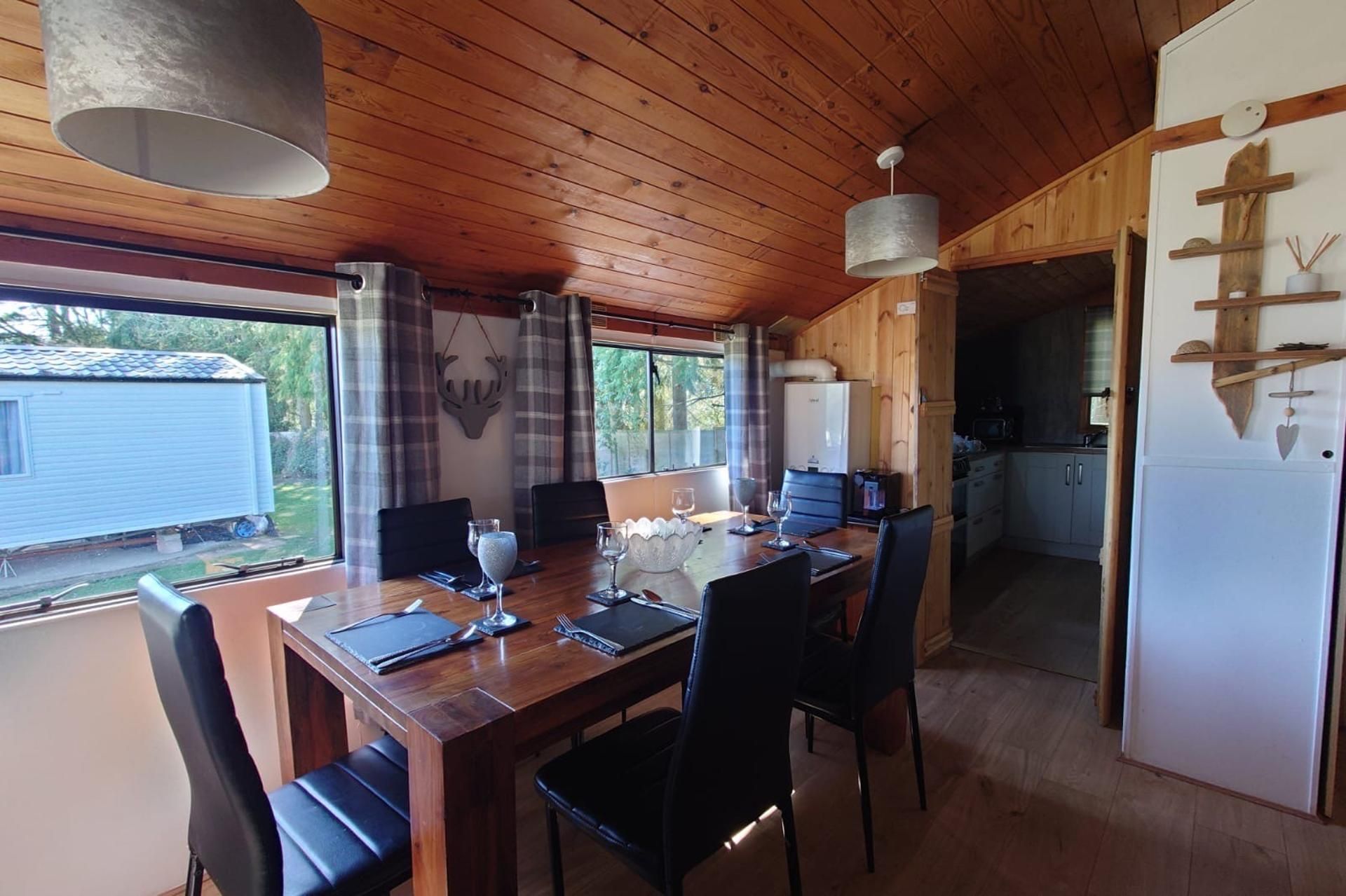 A wooden interior with a dining table set for four, a kitchen area, and a view of a mobile home outside.