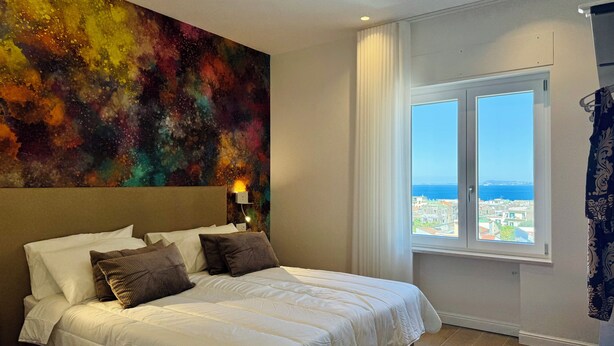 Une chambre d’hôtel avec un grand lit, un tableau abstrait aux couleurs vives accroché au mur, une fenêtre donnant sur la ville et un rideau à la fenêtre.