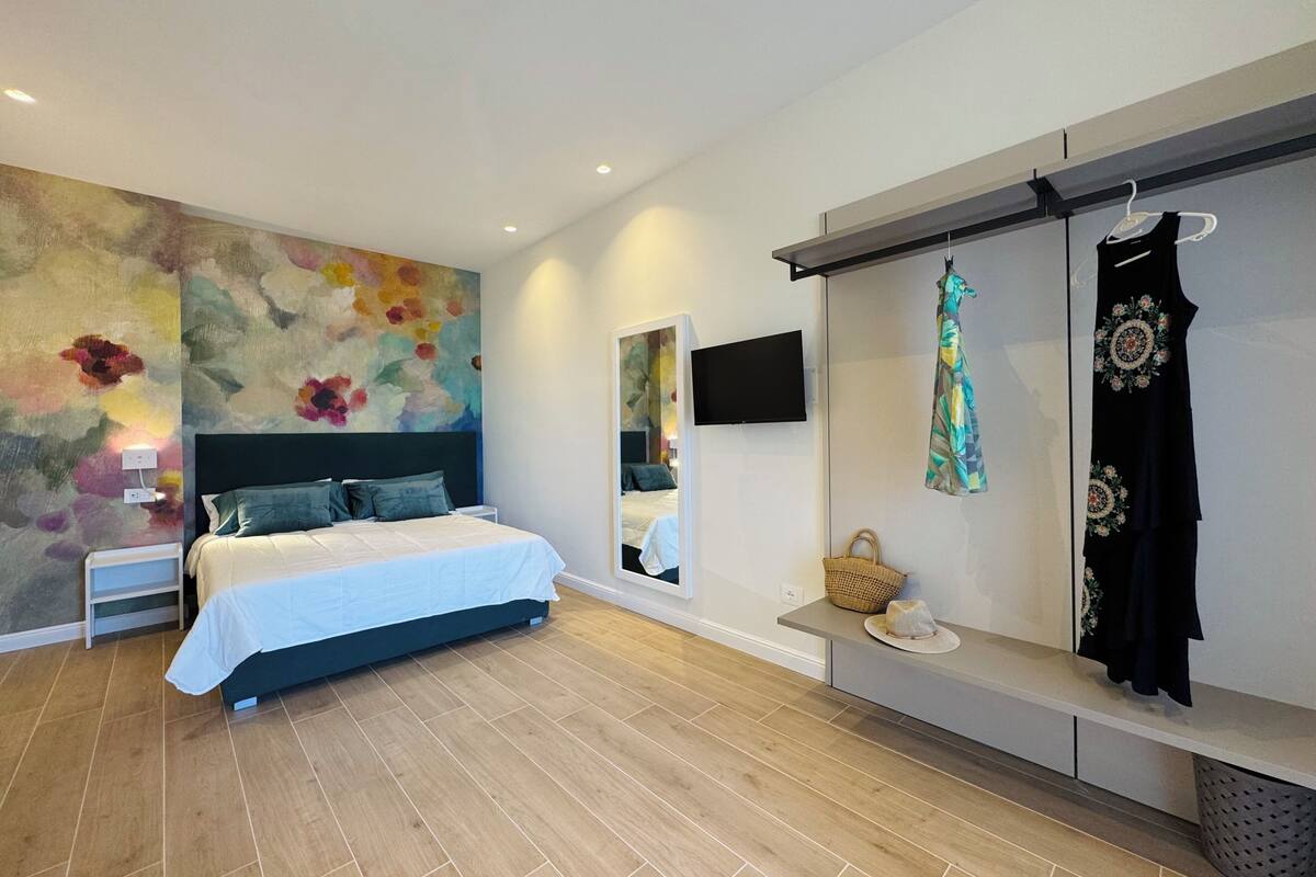 Un dormitorio moderno con una cama, un gran mural floral, un televisor y un armario con ropa colgada.