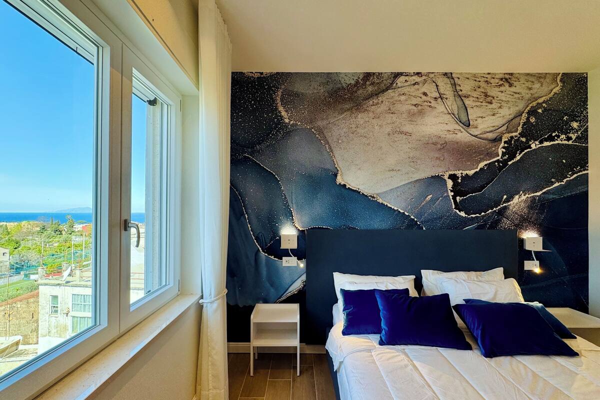 Un dormitorio con un ventanal grande, una cama con ropa de cama blanca y azul oscuro, y un mural en la pared.