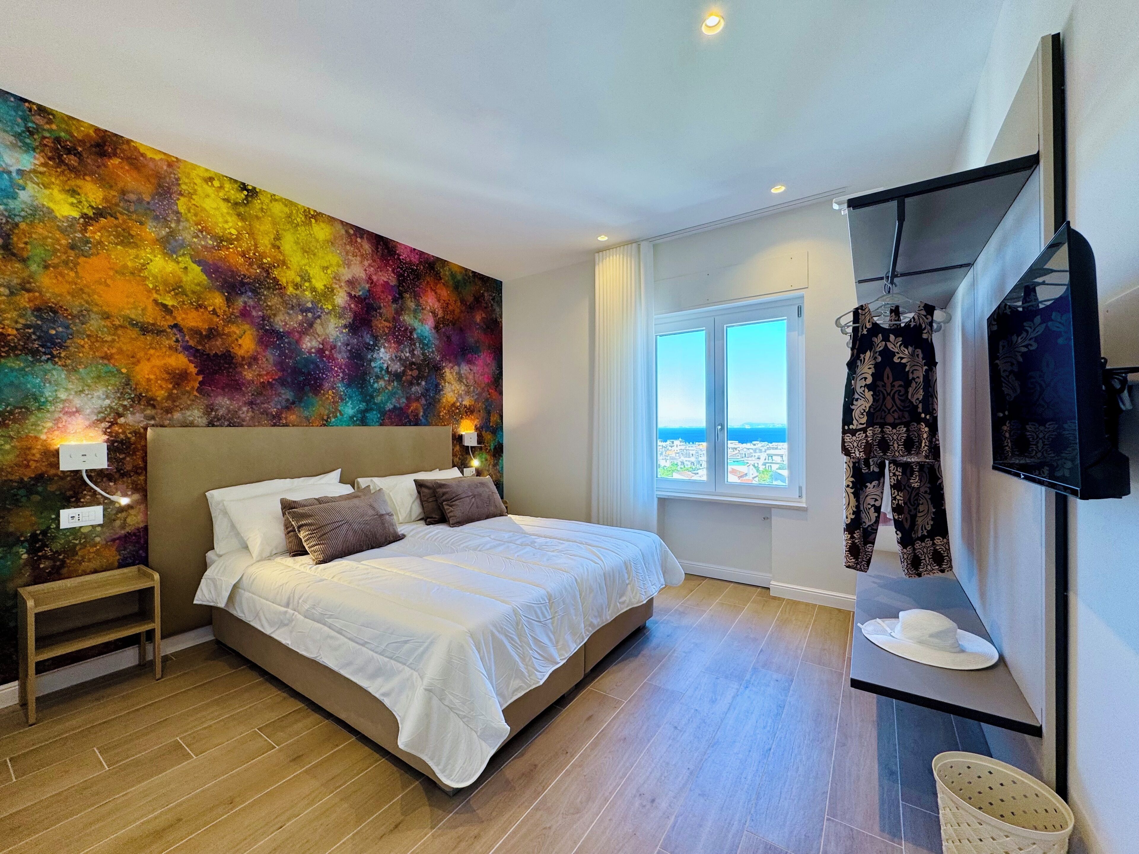 Une chambre dotée d’un grand lit, d’une fresque murale abstraite aux couleurs vives, d’une fenêtre offrant une belle vue et d’un téléviseur fixé au mur.