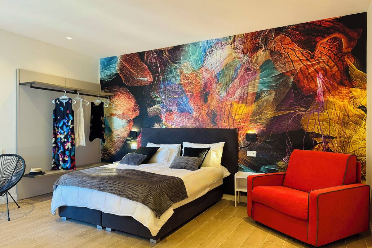 Un dormitorio moderno con una cama grande, un sillón rojo y un vibrante mural abstracto en la pared.