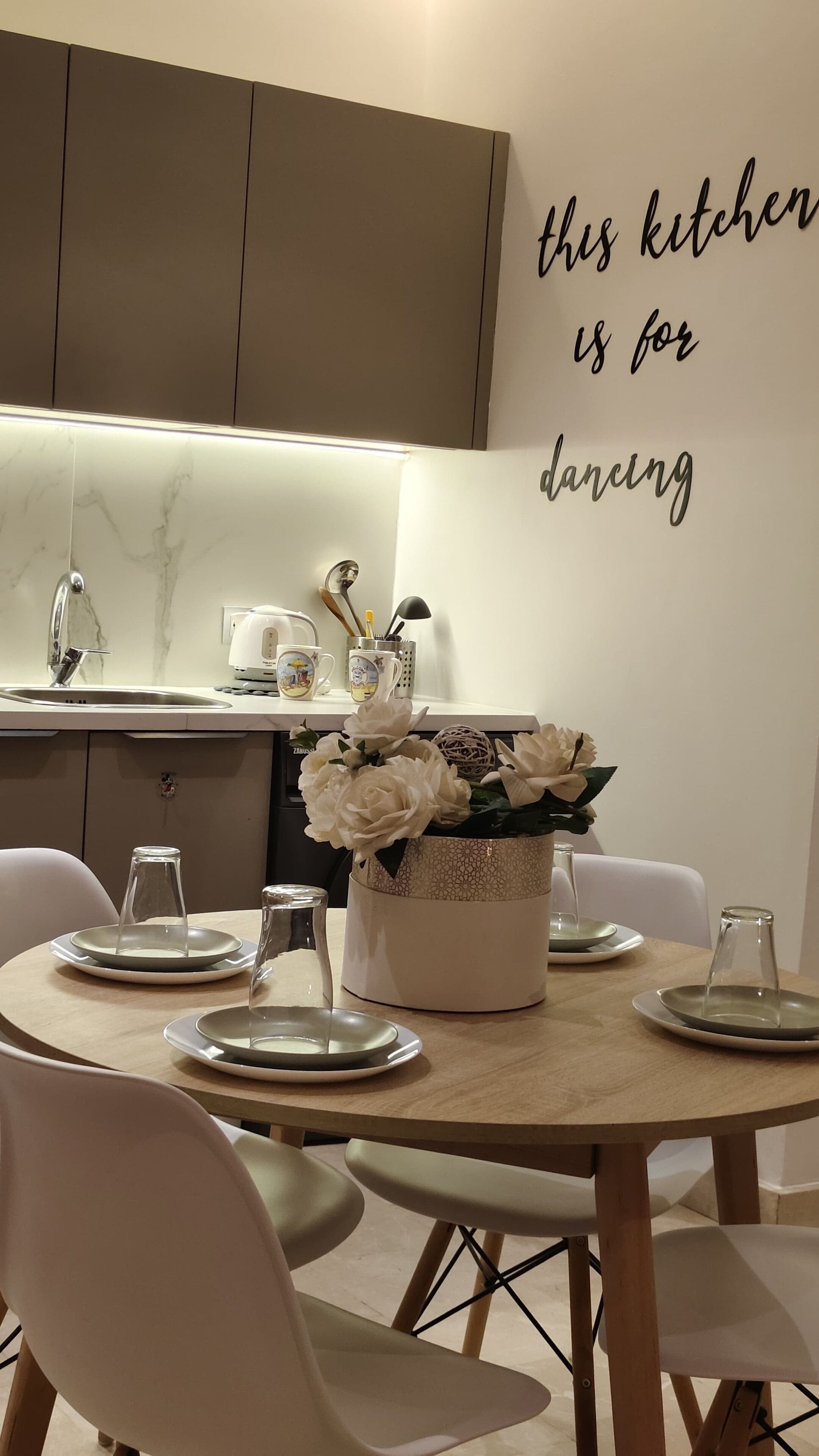 A modern kitchen with a marble backsplash, a dining table set with plates and a vase of flowers, and a wall sign that reads "this kitchen is for dancing".