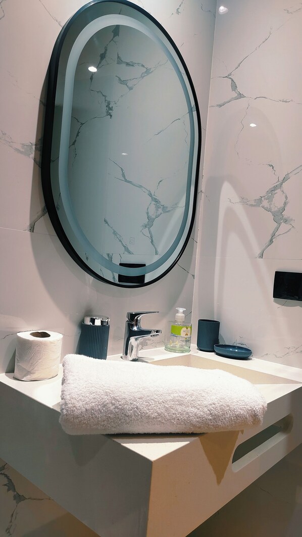 A bathroom with a marble backsplash, a round mirror, a white sink, a towel, a toilet paper roll, a soap dispenser, and a blue bowl.
