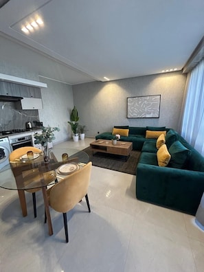 A modern living room with a green sofa, a glass dining table, and a kitchen area with a stove.
