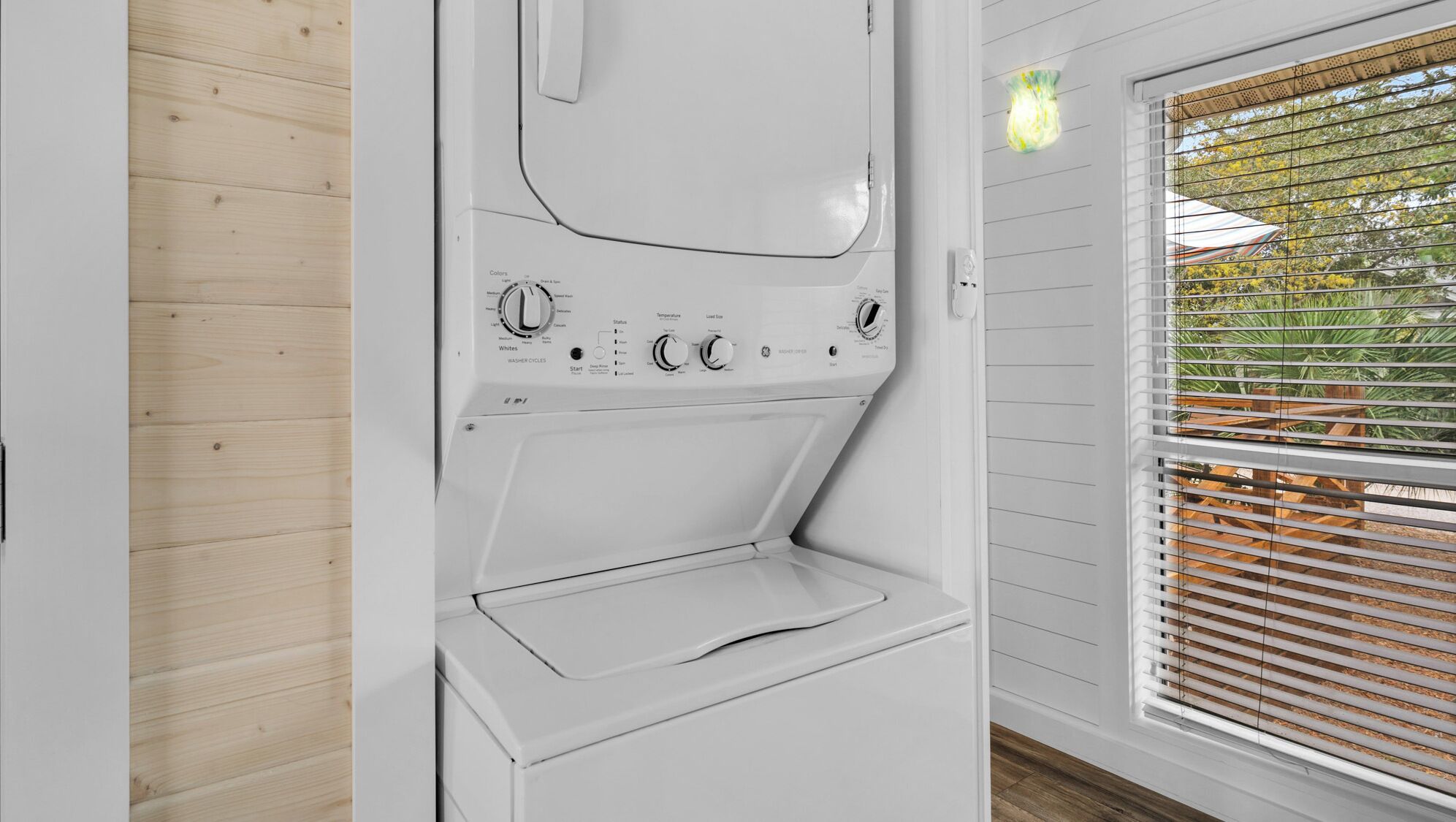 Una lavadora moderna y blanca con perillas de control y puerta transparente, ubicada en una habitación con piso de madera y una ventana con persianas.