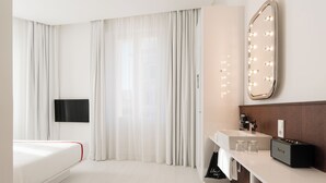 A modern hotel room with a bed, a TV, a vanity with a mirror, and a bathroom.