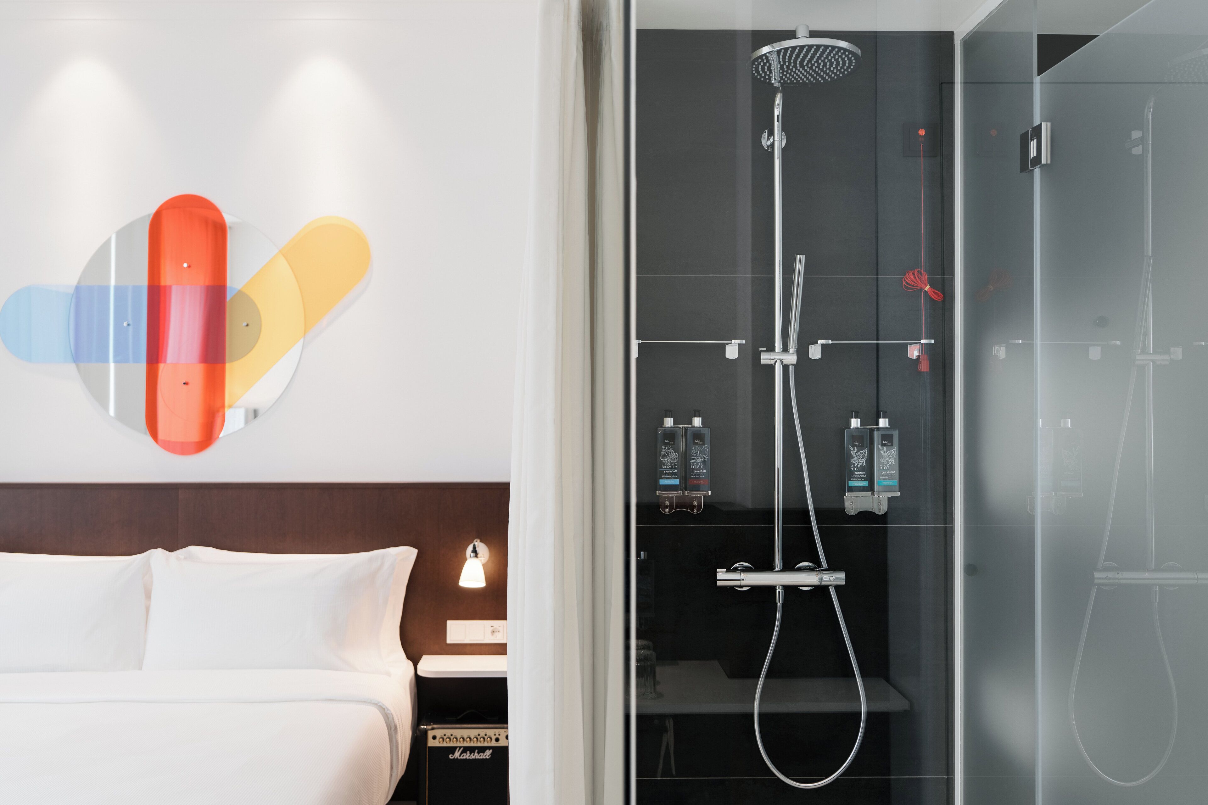 A modern hotel room with a bed, a glass shower enclosure, and a wall-mounted light fixture.
