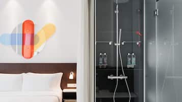 A modern hotel room with a bed, a glass shower enclosure, and a wall-mounted light fixture.