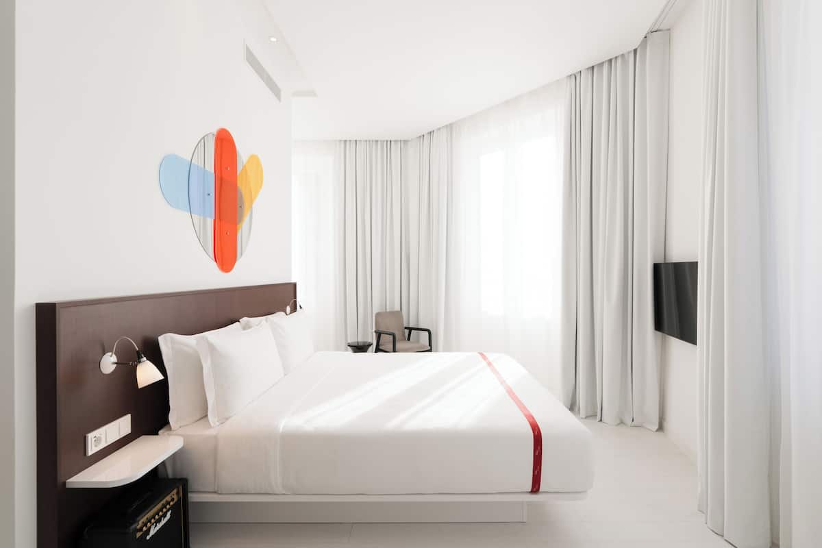 A modern hotel room with a large bed, a flat-screen TV, a desk with a lamp, and a chair.