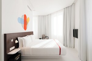 A modern hotel room with a large bed, a flat-screen TV, a desk with a lamp, and a chair.