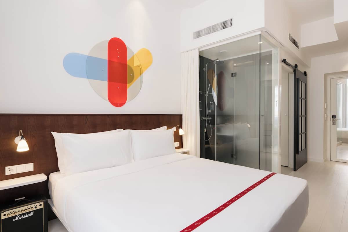 A modern hotel room with a large bed, a glass-enclosed shower, and a wall art piece.