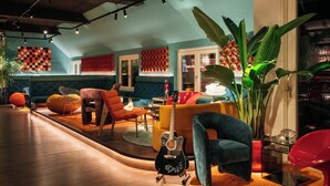 A modern lounge area with a curved wooden floor, vibrant seating options including orange and blue chairs, a guitar, and a large potted plant.