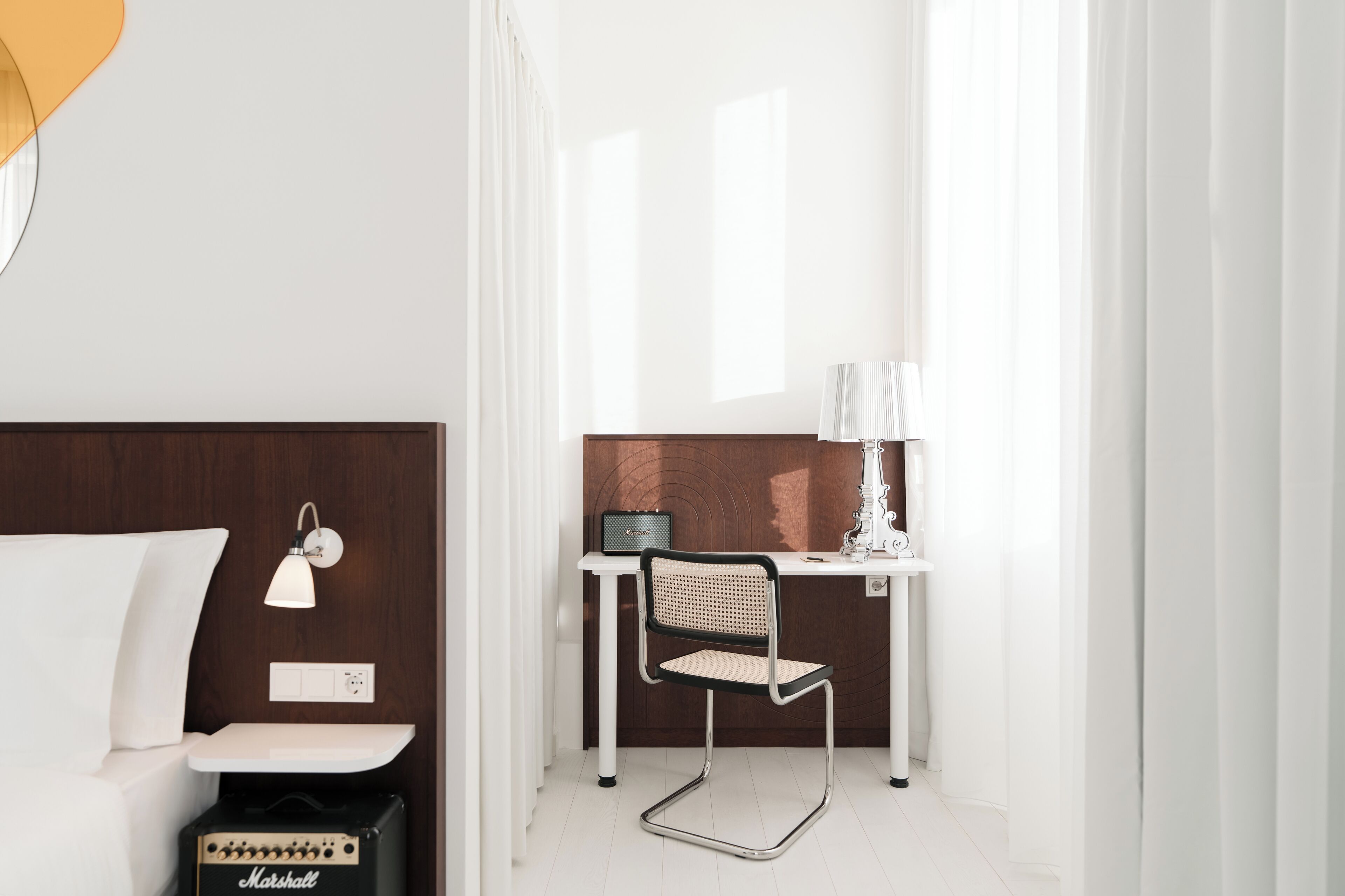 A modern hotel room with a bed, a desk with a chair, a lamp, and a Marshall amplifier.