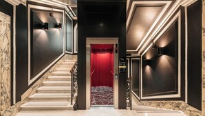 Un pasillo con suelo de mármol, una puerta de ascensor roja, paredes negras e iluminación en la pared.