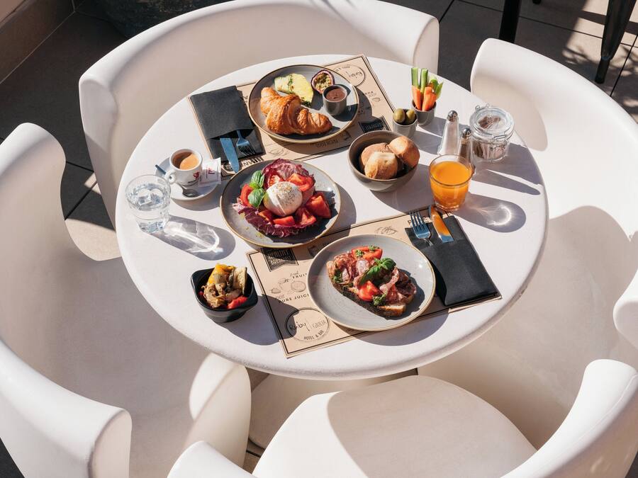 Mesa redonda con diversos productos para el desayuno, como croissants, fruta y pasteles, rodeada de sillas blancas.
