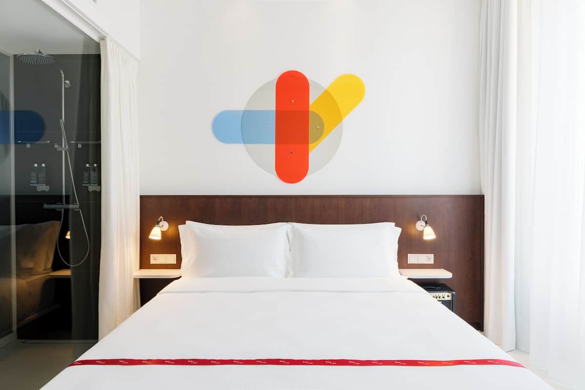 A hotel room with a bed, two bedside tables with lamps, a shower area, and a wall with a colorful heart design.