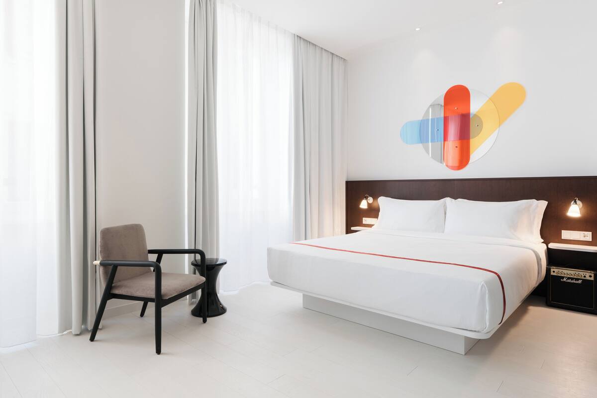 A modern hotel room with a large bed, a chair, a small table, and a nightstand with a lamp.