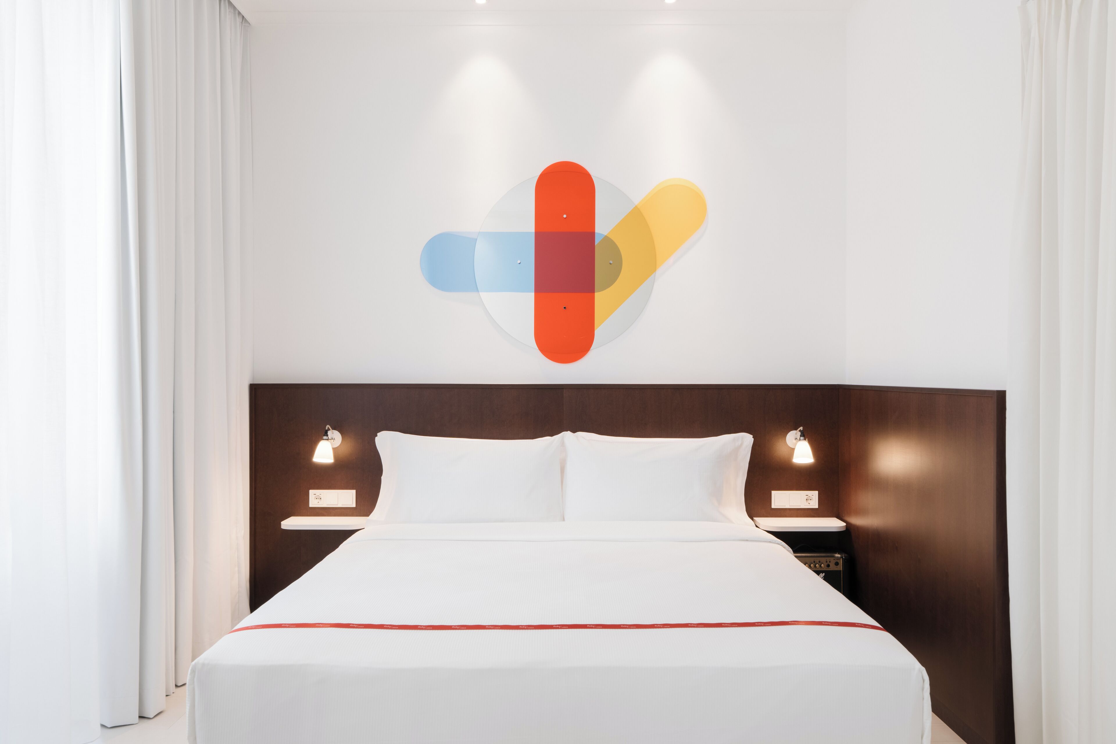 Ein modernes Hotelzimmer mit einem großen Bett, weißer Bettwäsche, einem Holzkopfende, Nachttischlampen und einem farbenfrohen Wandbild.