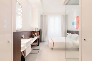 A modern hotel room with a bed, a desk, a mirror, and a bathroom.