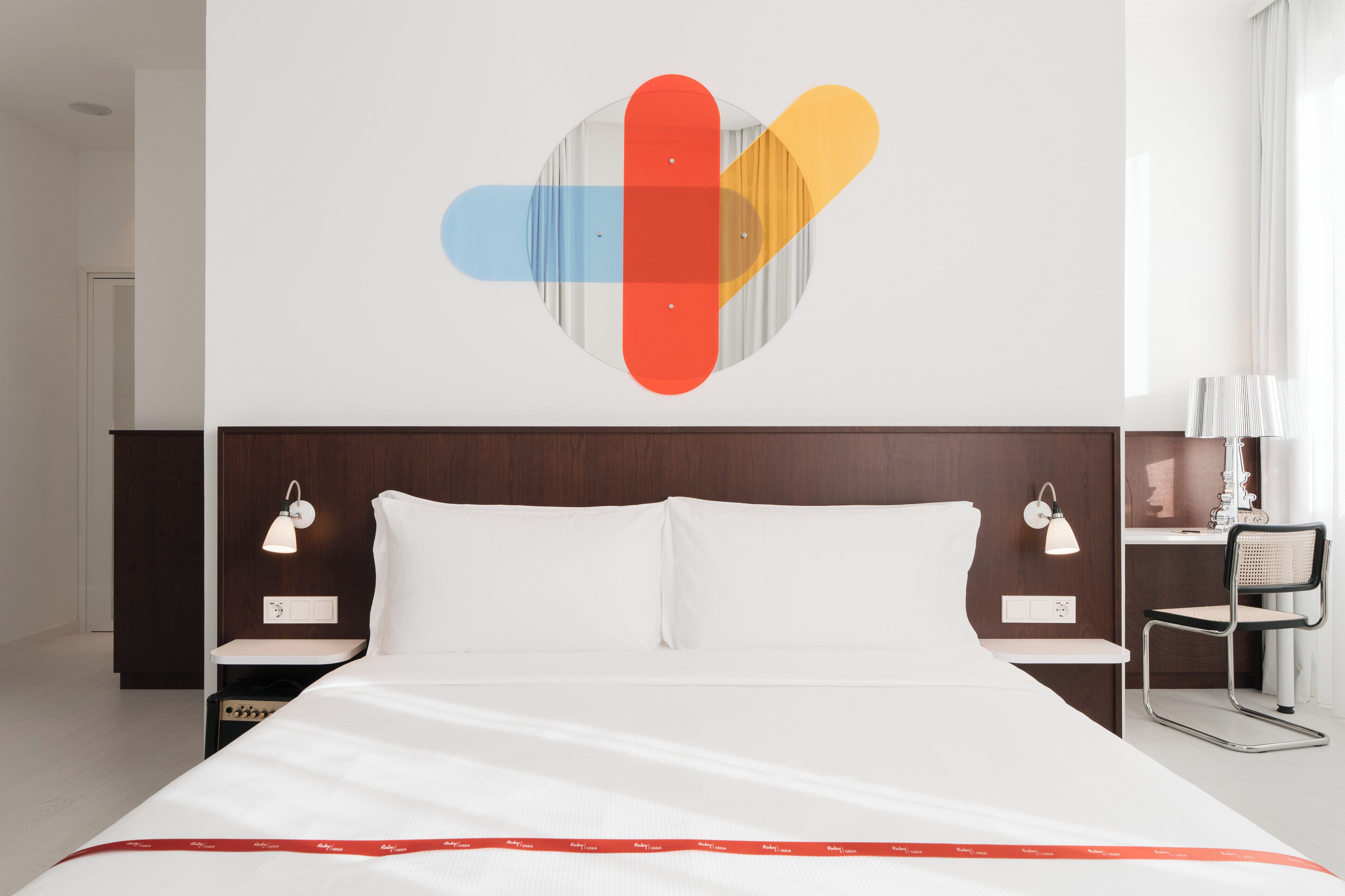 A modern hotel room with a large bed, two bedside tables with lamps, a wall-mounted artwork, and a chair.