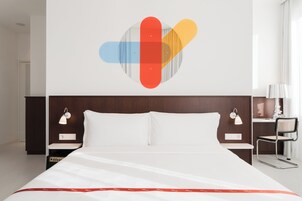 A modern hotel room with a large bed, two bedside tables with lamps, a wall-mounted artwork, and a chair.