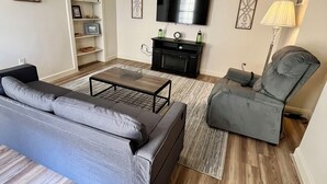 Una sala de estar moderna con un sofá y un sillón grises, una mesa de centro, un televisor de pantalla plana y una estantería decorativa.