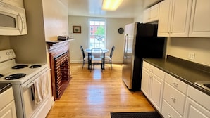 Una cocina con armarios blancos, una estufa y un refrigerador. Se ve una mesa de comedor con sillas al fondo.