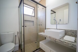 Um banheiro moderno com chuveiro em cabine de vidro, vaso sanitário branco e pia com bancada de granito e espelho acima.