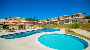 A swimming pool with a curved design, surrounded by a patio area with outdoor seating and a view of residential buildings. - Bz95 Cond. Viva Búzios Casa 4 quartos (Búzios)