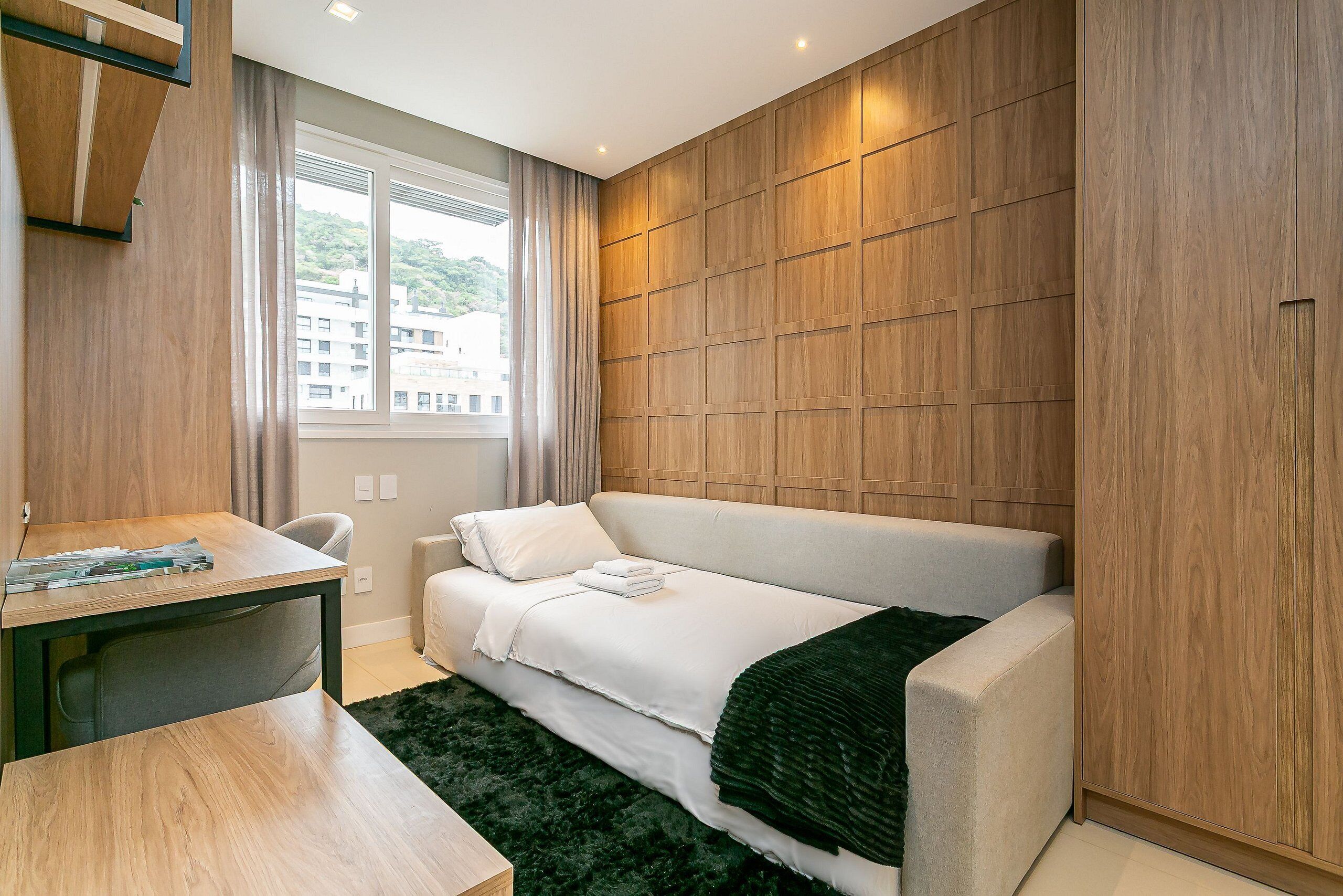 A modern bedroom with a wooden panel wall, a bed with white bedding, a desk with a chair, and a window with curtains.
