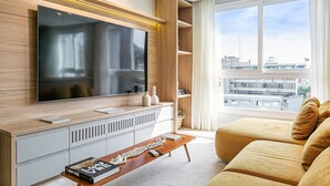 Moderni olohuone, jossa on suuri litteänäyttöinen televisio, puukalusteet, kirjoja sisältävä penkki ja suuret ikkunat, joista avautuu näkymä kaupunkiin.