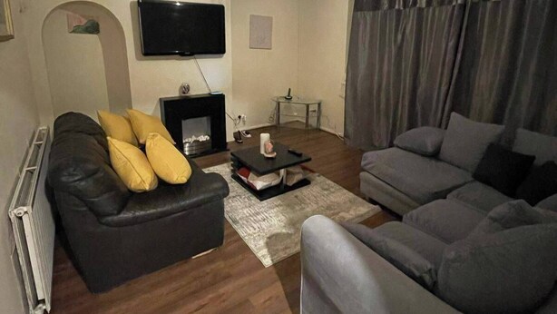 Ein Wohnzimmer mit schwarzem Ledersofa, einem grauen Sofa, einem am Wand montierten Flachbildfernseher, einem Kamin und einem Deckenventilator.